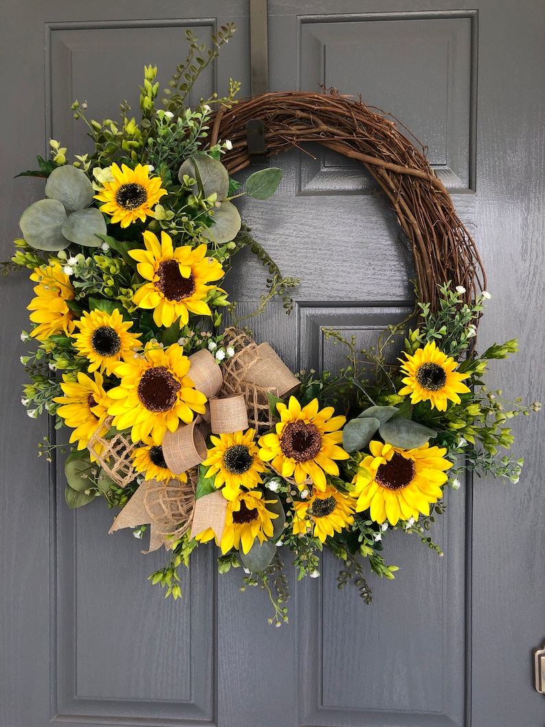 May include: A sunflower wreath with a burlap bow. The wreath is made of grapevine and features a variety of sunflowers, greenery, and small white flowers.