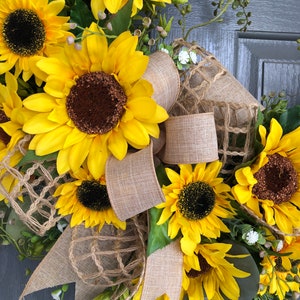 May include: A sunflower wreath with burlap ribbon and a brown mesh background. The wreath is decorated with yellow sunflowers and green leaves.