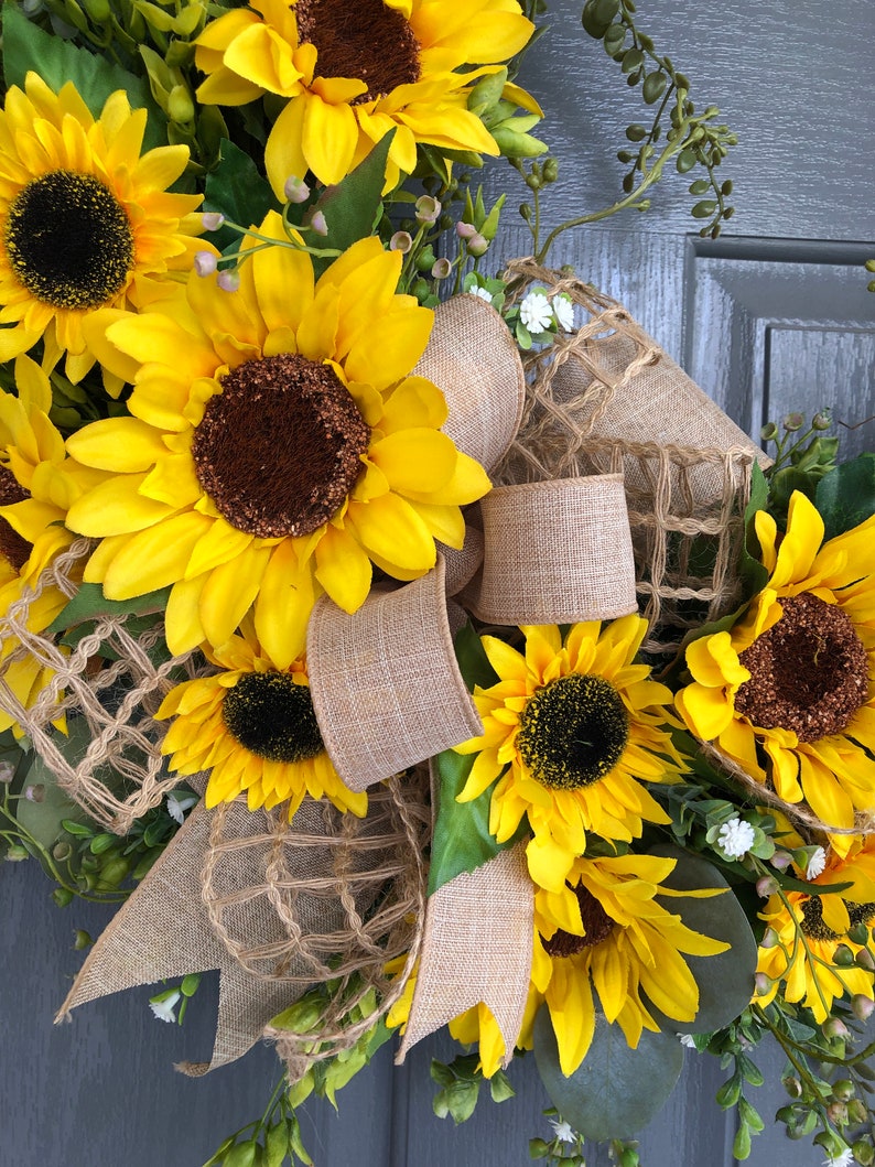 May include: A sunflower wreath with burlap ribbon and a brown mesh background. The wreath is decorated with yellow sunflowers and green leaves.