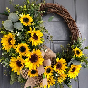 May include: A sunflower wreath with a burlap bow and greenery. The wreath is made of grapevine and features multiple yellow sunflowers with brown centers. The wreath is hanging on a gray door.
