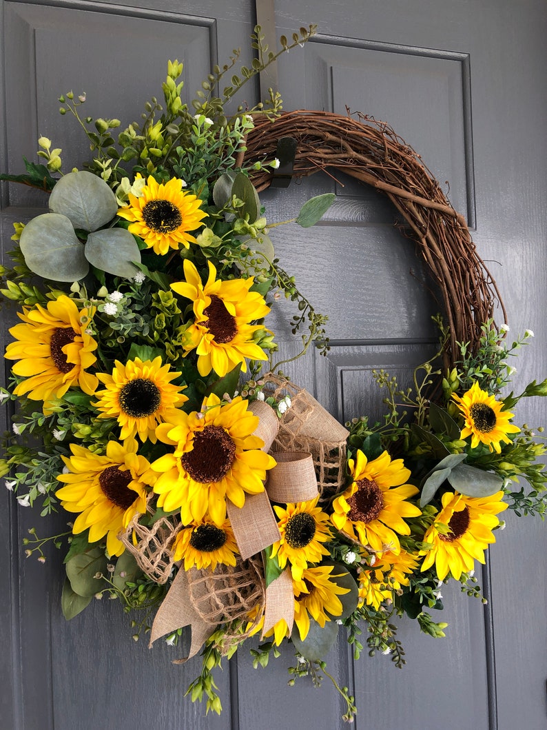 May include: A sunflower wreath with a burlap bow and greenery. The wreath is made of grapevine and features multiple yellow sunflowers with brown centers. The wreath is hanging on a gray door.