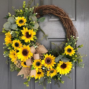May include: A sunflower wreath with a burlap bow. The wreath is made of grapevine and features a variety of sunflowers in different sizes. The wreath is perfect for adding a touch of summer to your home decor.
