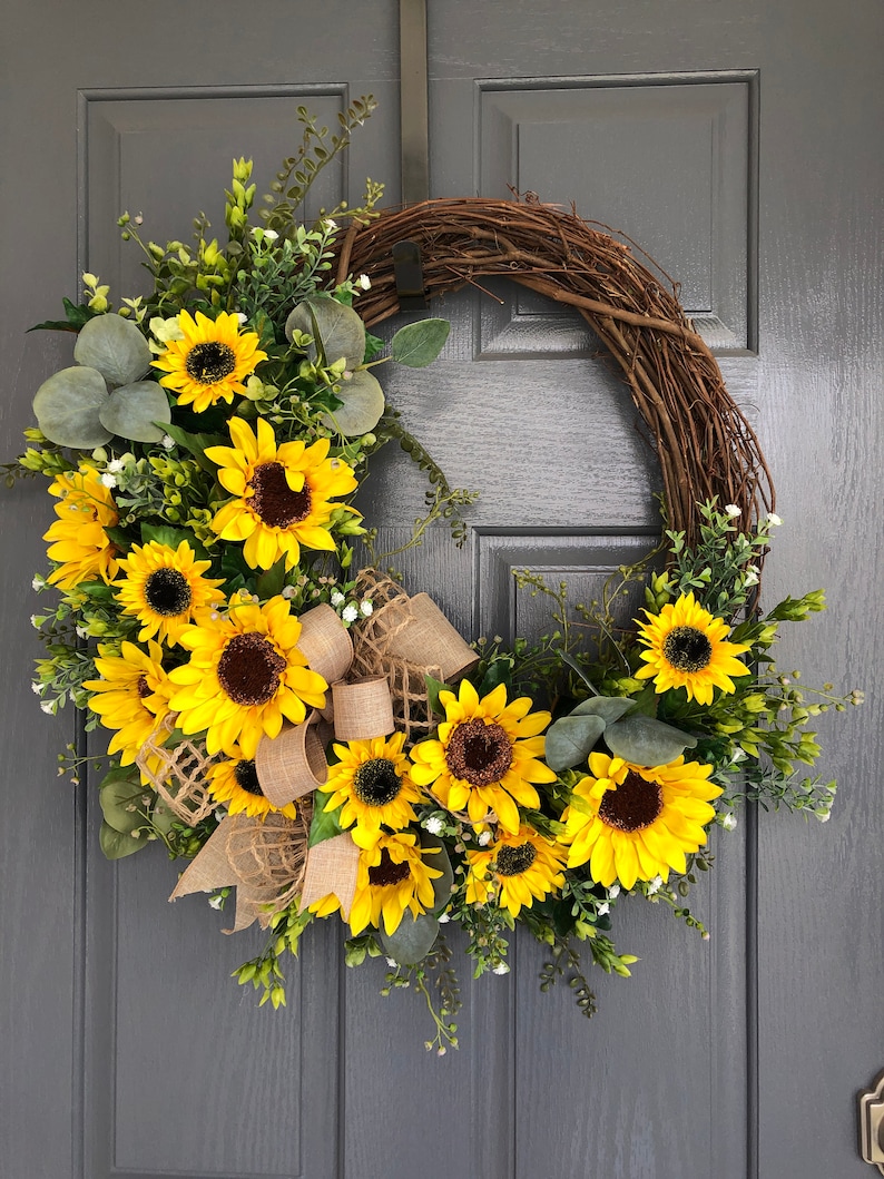 May include: A sunflower wreath with a burlap bow. The wreath is made of grapevine and features a variety of sunflowers in different sizes. The wreath is perfect for adding a touch of summer to your home decor.