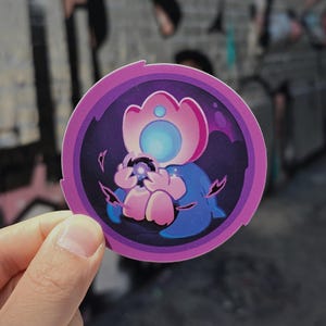 May include: A circular sticker featuring a stylised illustration of a small, blue, creature-like figure with a large, glowing pink and light blue head. The figure holds a dark, glowing orb, set against a deep purple background with a pink border.