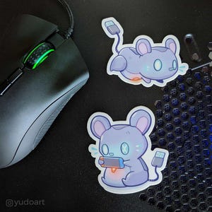 May include: Two stickers featuring a cartoon mouse holding a video game controller and a cartoon mouse with a USB cable. The mouse is light blue and gray with a white outline. The stickers are on a black surface.