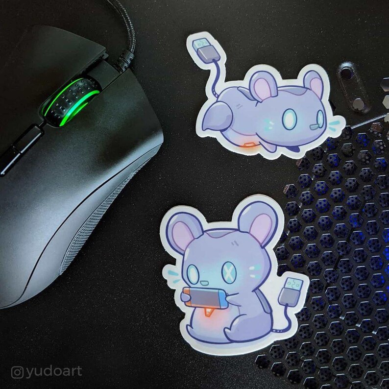 May include: Two stickers featuring a cartoon mouse holding a video game controller and a cartoon mouse with a USB cable. The mouse is light blue and gray with a white outline. The stickers are on a black surface.