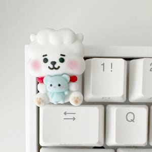 Könnte beinhalten: Eine weiße Tastatur mit einer weißen, flauschigen Figur, die eine kleine blaue Figur hält. Die Tastatur hat Tasten mit den Beschriftungen 1!, 2@, 3#, Q, W, E, A, S, D, Z, X, Ctrl, Alt und eine Windows-Taste.