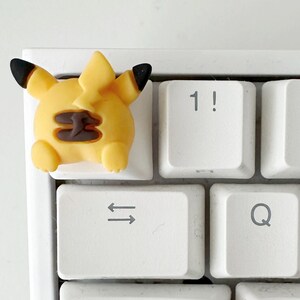 May include: A white keyboard with a yellow and brown Pikachu character keycap on the '1!' key. The keyboard has the standard layout with keys for numbers, letters, and symbols.