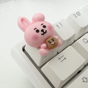 Puede incluir: Un personaje de conejo rosa con ojos marrones y una nariz marrón sosteniendo un personaje de oso marrón con una nariz y ojos marrones. Los personajes están en un teclado blanco.