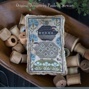 May include: A cross-stitch sampler featuring two sheep, one black and one gray, with floral accents. The sampler is displayed in a wooden bowl with spools of thread. The text "Plum Street Samplers" and "Snort Stack" are visible.