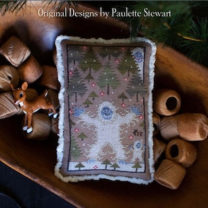May include: A cross-stitch sampler with a winter theme, featuring a yeti and trees, is displayed in a wooden bowl. The sampler is surrounded by brown thread spools and a small deer figurine. The text "Abominable Bundle" is at the bottom.