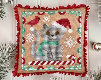 May include: A square, tan Christmas cushion with a cross-stitch design. The design features a grey cat wearing a Santa hat and green scarf, a red bird, and snowflakes. The cushion has a red ruffled border and candy cane accents.