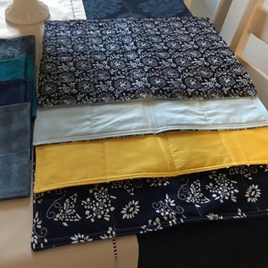 Pode incluir: Uma coleção de individuais retangulares em várias cores e padrões. Inclui cores sólidas como amarelo, azul e verde-azulado, bem como designs estampados com motivos florais brancos sobre um fundo azul marinho.