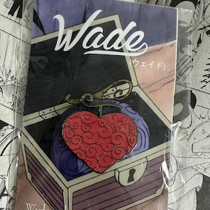 May include: A pin featuring a red heart-shaped fruit with a green stem inside a wooden treasure chest. The pin has the word "Wade" in white script above the chest. The website "Wvdeandco.com" is also visible.