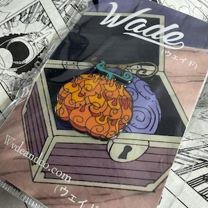 May include: A pin featuring a stylized orange and yellow fruit and a purple fruit inside a treasure chest design. The pin is on a backing card with the name "Wade" and the website "Wvdeandco.com". The background includes manga panels.