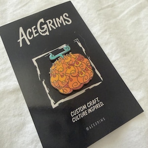 May include: A metal enamel pin shaped like a stylised orange and yellow fruit with a teal stem, mounted on a black display card. The card features the white text ACEGRIMS at the top and CUSTOM CRAFT. CULTURE INSPIRED. at the bottom right.
