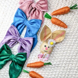 May include: Four velvet hair bows in pink, blue, purple, and green, arranged in a stack. Three brown twine carrots and a burlap bunny with a daisy are on a white knitted surface.