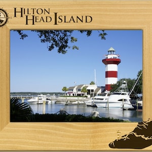 May include: A wooden picture frame with a compass and the text "HILTON HEAD ISLAND" engraved on the top. The frame contains a photo of a red and white striped lighthouse with boats in the harbor.