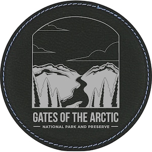 May include: Black round leather coaster with a stitched edge. The coaster features a white graphic of a mountain range with a river flowing through it. The text "Gates of the Arctic - National Park and Preserve" is printed below the graphic.