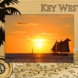 May include: A wooden picture frame with a sunset scene of a sailboat on the ocean. The frame has a map of the Florida Keys with the text "KEY WEST" at the top.