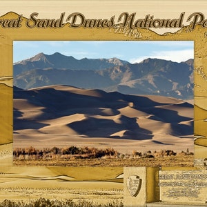 May include: A scenic view of the Great Sand Dunes National Park. The image features rolling sand dunes in the foreground, with mountains and a clear blue sky in the background. The park's name is displayed in a decorative frame.