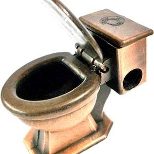 May include: A miniature bronze-toned metal toilet figurine featuring a hinged lid that opens upward. The base is rectangular, and the tank section includes a circular opening on the side. The surface has a weathered, metallic finish with dark accents.