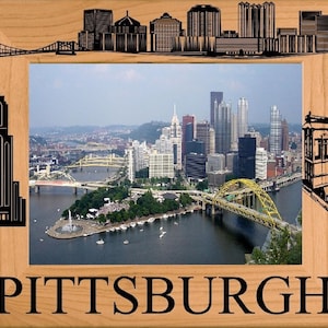 May include: A wooden picture frame with a black silhouette of the Pittsburgh skyline on the top and a black silhouette of a building on the left. The frame has a photo of the Pittsburgh skyline with a river and bridges in the foreground. The frame has the word "PITTSBURGH" in black letters on the bottom.