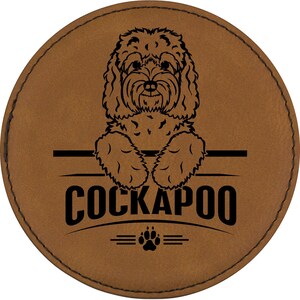 May include: A round, brown leather coaster with a black engraved Cockapoo dog design. The design includes the dog's face and paws, the word "COCKAPOO", and a paw print symbol.