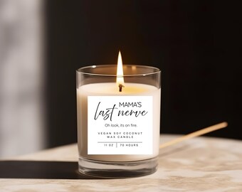 May include: A lit candle in a clear glass jar. The candle has a white label with the text "MAMA'S last nerve" and "Oh look, its on fire." The label also states "VEGAN SOY COCONUT WAX CANDLE" and "11 oz | 70 HOURS."