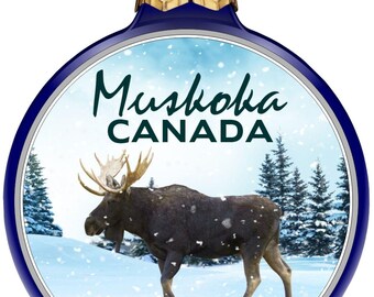 May include: A Christmas ornament with a blue border and a gold top. The ornament features a moose in a snowy landscape with the text "Muskoka CANADA". The moose is brown with large antlers.