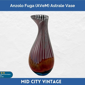 May include: Anzolo Fuga (AveM) Astrale Vase. A tall, decorative glass vase with a unique shape. The vase features vertical stripes in shades of red, white, and black. The base is a mix of brown and blue. The text "Mid City Vintage" is at the bottom.