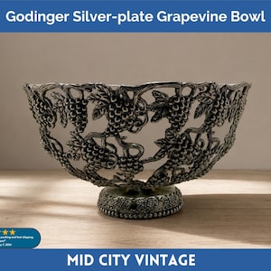May include: Godinger silver-plate grapevine bowl. The bowl features a detailed grapevine design with clusters of grapes and leaves. It has a pedestal base and a decorative rim. The bowl is silver-colored and sits on a wooden surface.