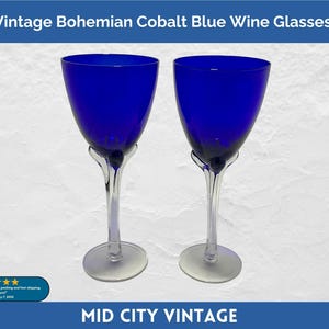 May include: Two vintage cobalt blue wine glasses. The glasses have a clear stem and base, with a deep blue bowl. The text "Vintage Bohemian Cobalt Blue Wine Glasses" is at the top of the image. The glasses are on a white background.