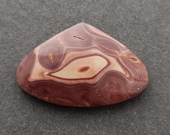 Op de afbeelding: Een driehoekige cabochon van rood, wit en bruin gebande agaat. De steen heeft een draaiende structuur en is gepolijst.