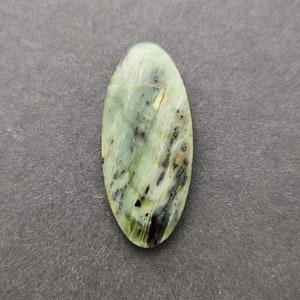 Könnte beinhalten: Ein grüner und schwarzer ovaler Cabochon-Edelstein mit goldenen Flocken. Der Stein hat ein wirbelndes Muster und ist vor einem grauen Hintergrund platziert.