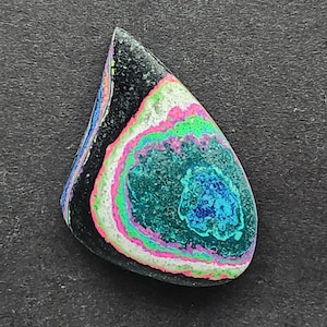 May include: A teardrop-shaped cabochon of black, green, blue, pink, and white opal. The opal has a swirling, layered pattern.