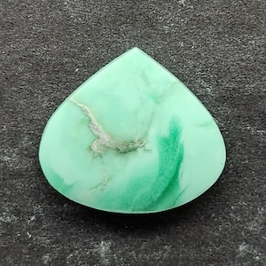 Puede incluir: Una gema en forma de lágrima, de color verde claro y aspecto veteado. La piedra presenta un patrón veteado más claro, casi blanco, en su superficie, con vetas verdes más oscuras. La gema se encuentra sobre un fondo gris oscuro.