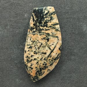 May include: A polished gemstone with a teardrop shape. The stone features a mix of beige, black, and green colors. The pattern includes black, needle-like inclusions and cross-like markings. The gemstone is set against a dark gray background.
