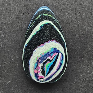 May include: A teardrop-shaped cabochon with a swirling pattern of black, white, blue, green, and pink. The cabochon is a natural stone with a unique and colorful design.