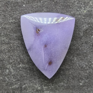 May include: A triangular-shaped lavender gemstone with a smooth, polished surface. The stone features a soft, opaque purple hue with three distinct, small, dark brown inclusions scattered across its face, creating a unique natural pattern.