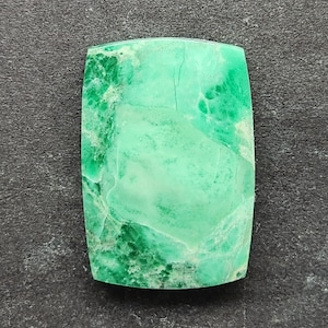 Puede incluir: Una piedra preciosa rectangular con esquinas redondeadas, que muestra una mezcla de tonos verdes claros y oscuros. La piedra tiene un aspecto veteado con vetas visibles, lo que sugiere un origen natural. La superficie pulida refleja la luz.