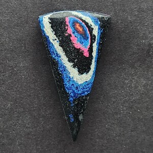 May include: A black, blue, white, and pink cabochon with a swirling pattern. The cabochon is shaped like a triangle with a rounded top.
