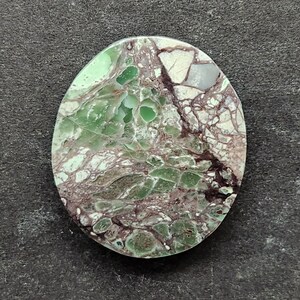 Puede incluir: Gema ovalada con un patrón único de tonos verdes, blancos y granates. La superficie de la piedra muestra un efecto veteado, con vetas granates que cruzan los colores más claros. La apariencia general es orgánica y natural, sugiriendo una piedra pulida.