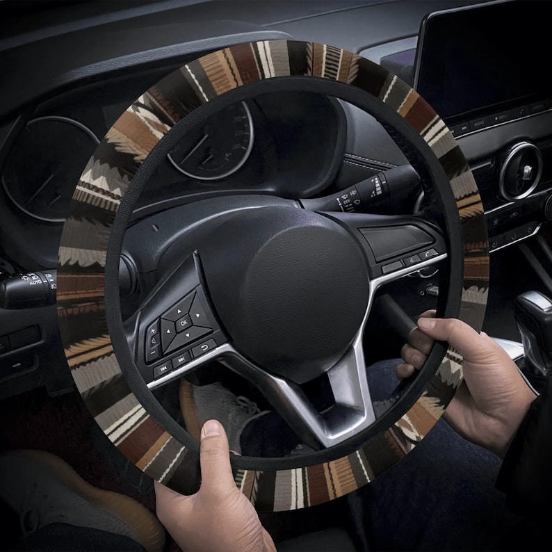 May include: A patterned steering wheel cover with a black border. The cover features a design of brown, tan, and white stripes and shapes. The cover is on a black steering wheel in a car.