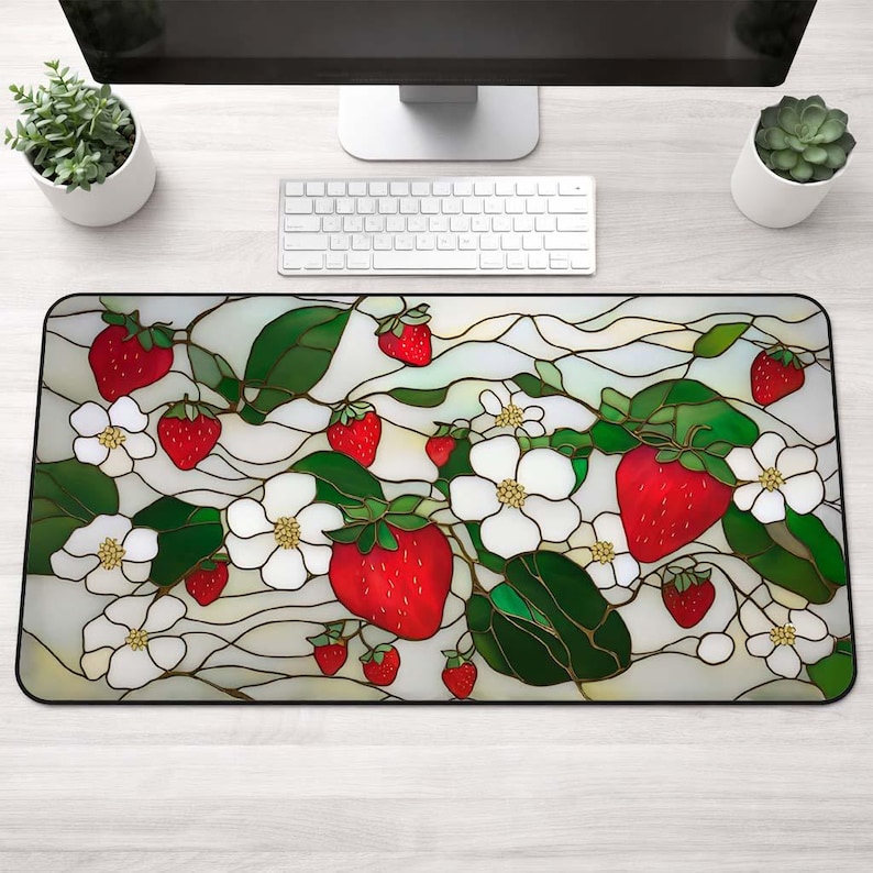 May include: A rectangular desk mat featuring a stained-glass design of strawberries, white blossoms, and green leaves. The mat has a black border and a light background. The strawberries are red, and the flowers are white.