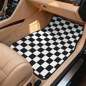 May include: A rectangular car floor mat featuring a high-contrast black and white checkered pattern. The mat is positioned in the front passenger footwell of a vehicle with tan interior upholstery and a black dashboard console.