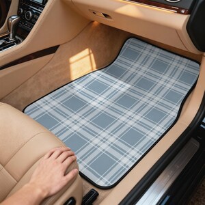 May include: A rectangular car floor mat featuring a light blue and white plaid pattern with a black border. The mat is positioned in the passenger footwell of a vehicle with tan interior upholstery and a dark center console.
