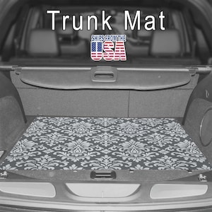 May include: A rectangular trunk mat featuring a repeating white damask pattern on a dark gray background, installed flat on the floor of a vehicle cargo area. The text "Trunk Mat" appears at the top, with a "SHIPS FROM THE USA" flag graphic.