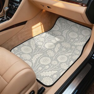 May include: A rectangular car floor mat featuring a light gray background with a repeating pattern of white dandelion flowers, buds, and jagged leaves. The mat has a black border and is positioned in the front passenger footwell of a vehicle.