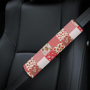 May include: A rectangular seat belt cover featuring a patchwork design of red and white patterns. The fabric displays alternating squares of red gingham, white with red polka dots, and white with red strawberry and small flower illustrations.